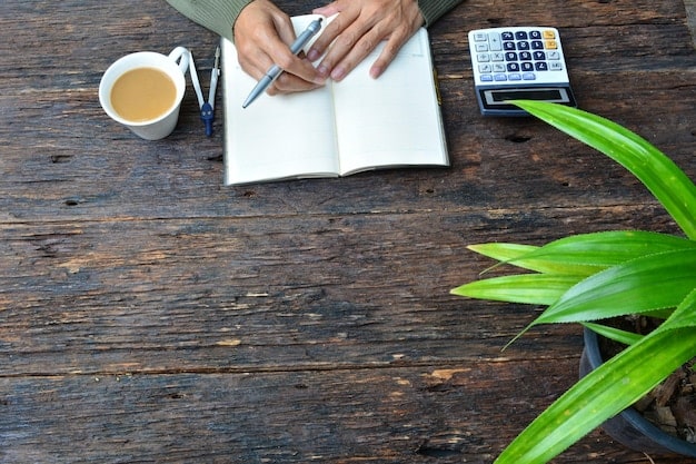 A close-up shot of a person writing down financial goals in a notebook. The notebook is open on a desk with a calculator and other financial documents surrounding it.